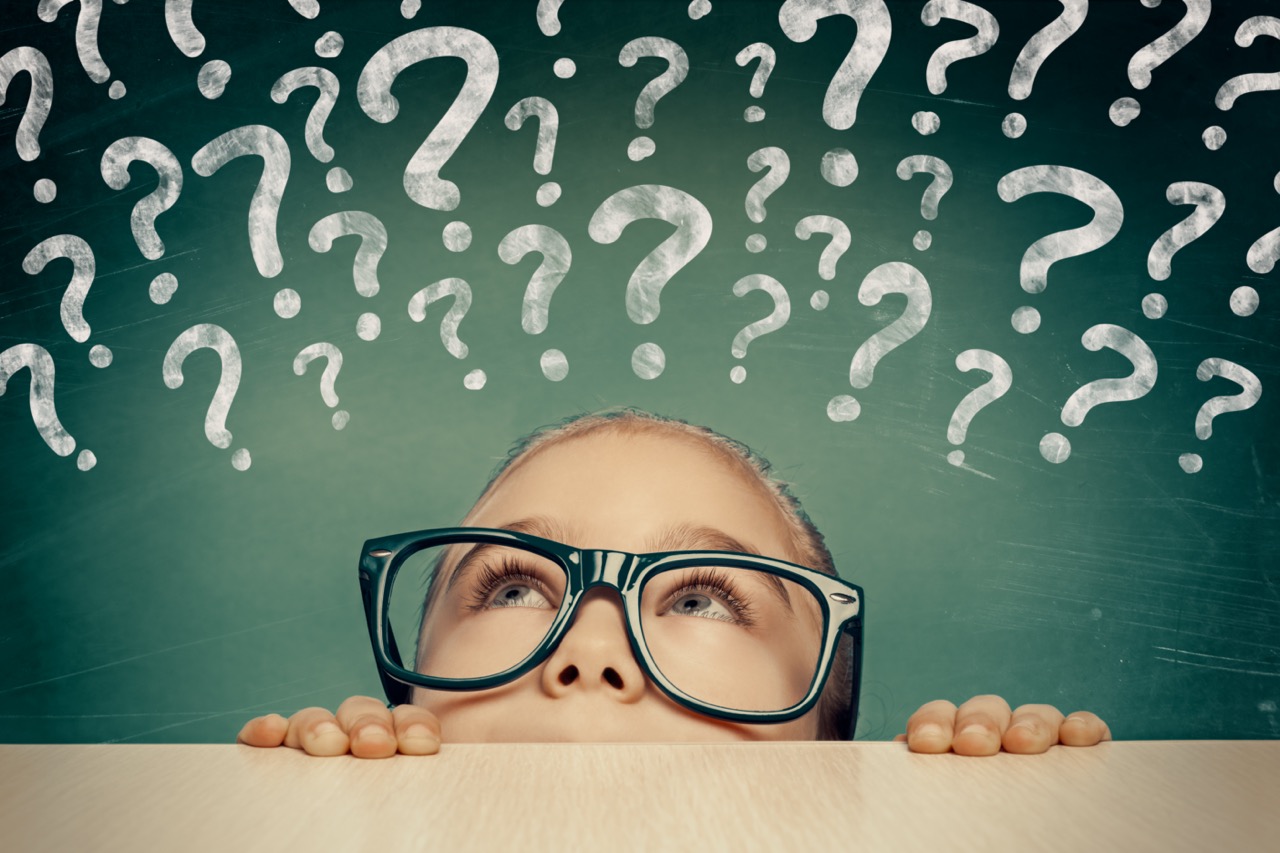 beautiful cute little girl looking at question marks above her head.  confused little student in front of chalkboard. 