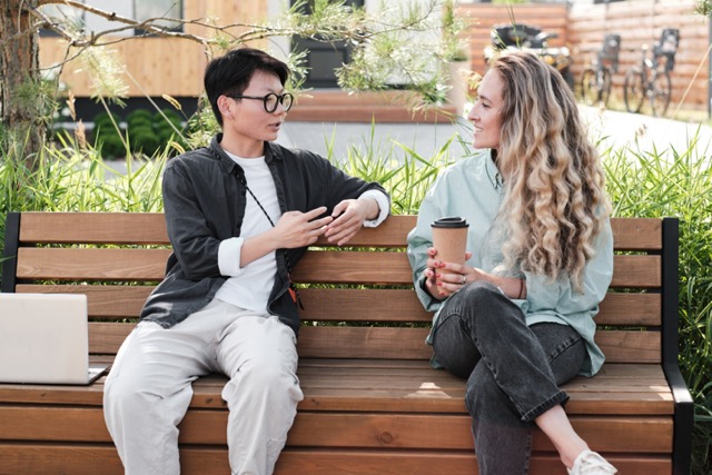 Horizontal medium long portrait shot of two multi-ethnic young women wearing casual clothes sitting together on bench outdoors drinking coffee and chatting during break